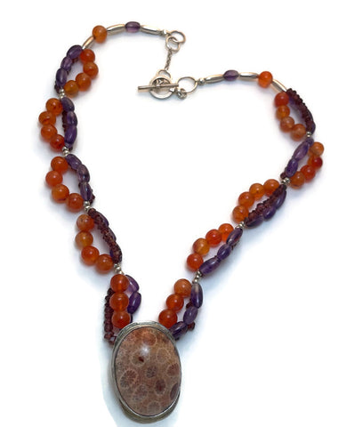 A very unique necklace with multi strands of opaque orange agates, tubular shaped amethysts and small reddish purple garnets interspersed with sterling silver beads.  The focal point is a piece of fossilized coral set in sterling silver.  The coral has colors of pinkish orange and red.  One-of-a-kind.  