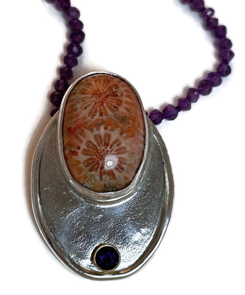 A zoom shot of the pendant.  A beautiful orange -toned fossilized coral with a floral design from the fossil occupant is accented with a 14KY gold set deep purple amethyst. The pendant is all sterling silver and strung on pearl knotted purple amethysts with orange sunstones that shimmer in the sunlight. One-of-kind.  