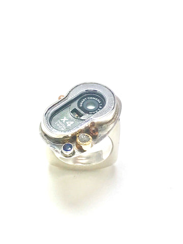 A ring made out of a cell phone camera ring.  The camera is accented with a blue sapphire and a diamond and one small gold ball.